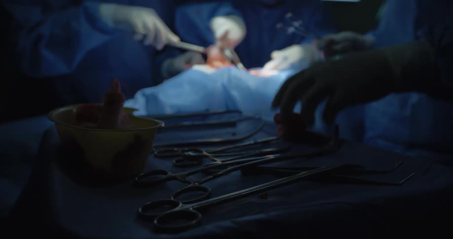 Surgical instruments are laid out on a sterile drape in the foreground while surgeons dressed in blue scrubs and gloves perform a procedure under focused lighting in the background