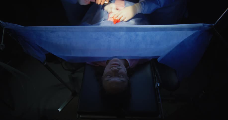 Woman lying asleep on a surgical bed and people with gloves operating on her abdominal area