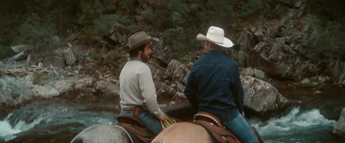 Two cowboys are riding horses along a riverbank with one wearing a dark hat and the other wearing a white cowboy hat both facing the flowing water and rocky terrain