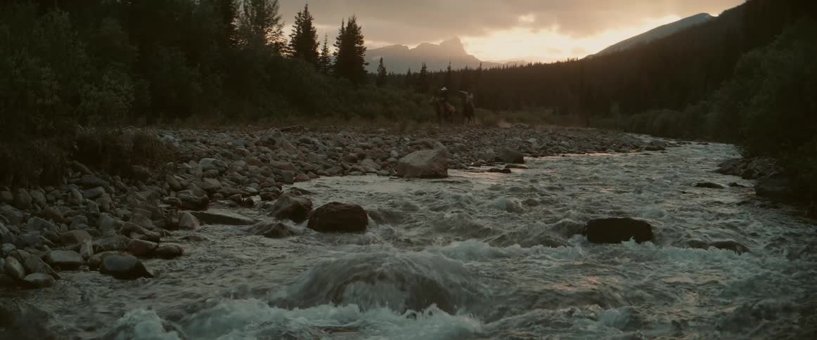 Rushing river during a cloudy dusk sky two people riding fast along the riverbank on horses there is a distant mountain