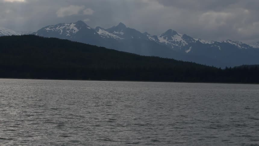 Dark water meets forested hills and snowy peaks beneath cloudy skies
