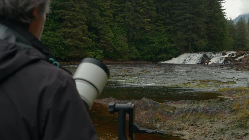 A photographer aims a telephoto lens at a waterfall scene in a lush forest setting