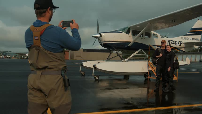 Man takes a photo of two people posing beside a small plane on a wet tarmac