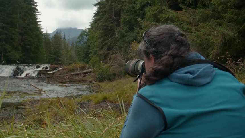 A person photographs a waterfall in a forest from a grassy riverbank