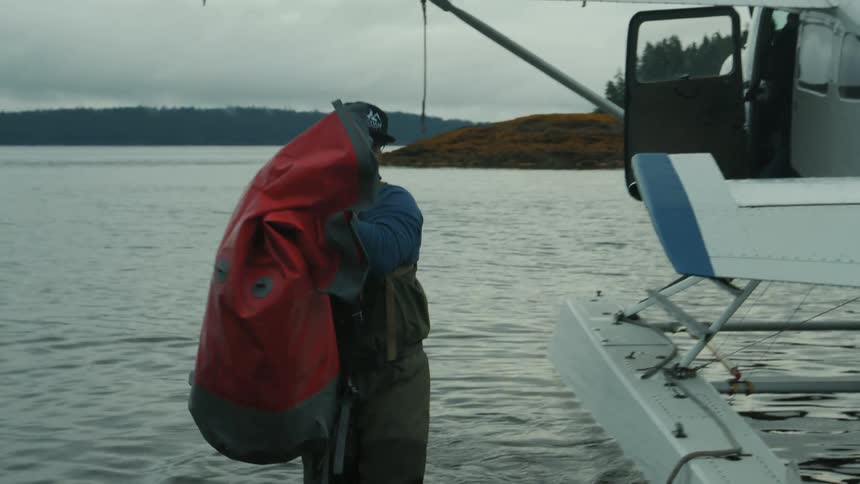 A man wearing a hat and glasses carries a bag from a seaplane as another person in the plane hands it to him he walks from the plane in the lake to the shore drops the bag then walks back into the lake to retrieve another bag on a cloudy day