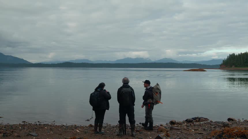 Three hikers stand at a lakeshore gazing at the distant mountains