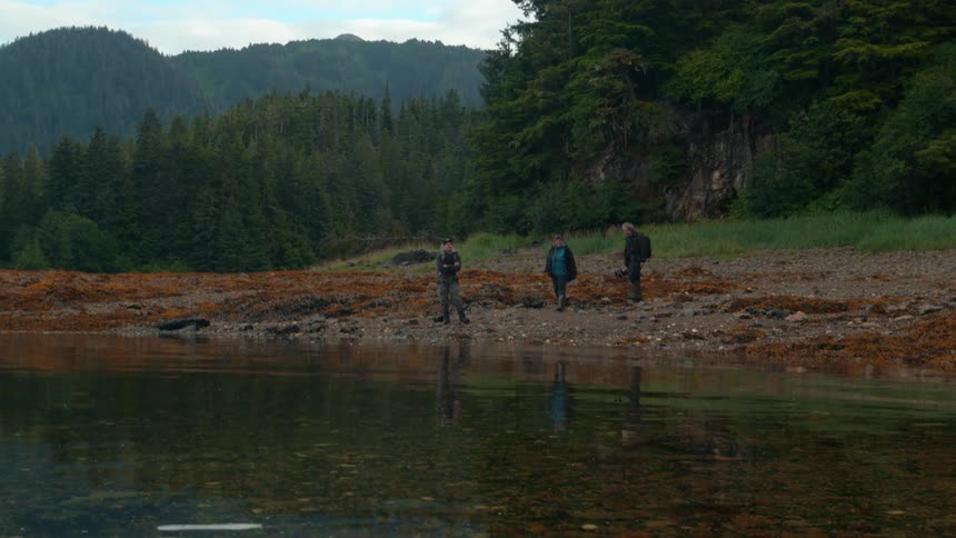 Three hikers walk along a rocky forest shoreline by calm water