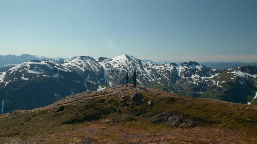 A mountain trail leads to two people standing on the peak staring at another snowcovered mountain in the distance