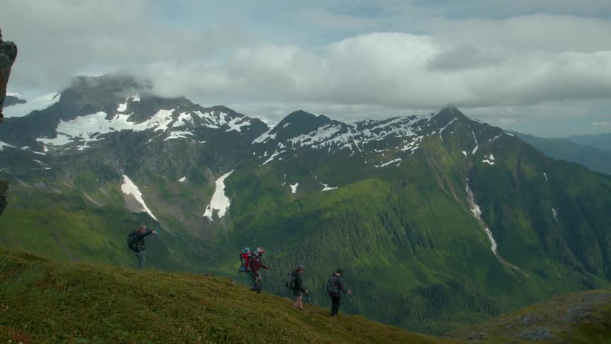 Hikers trek along a grassy ridge with snowcapped mountains in the background