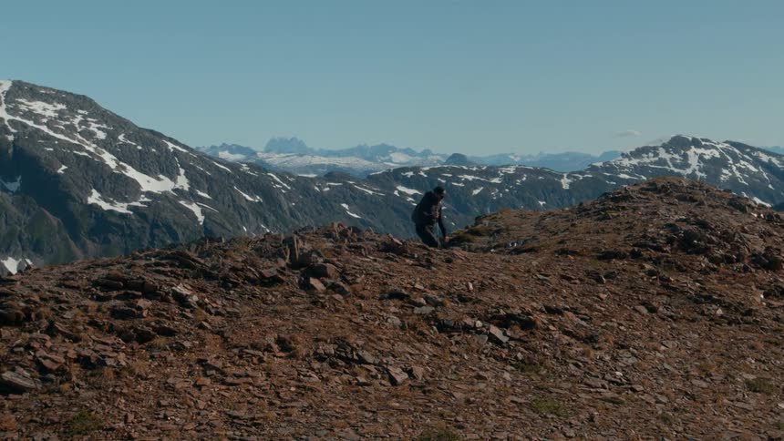 Two hikers trek a rocky ridge with snowy peaks behind