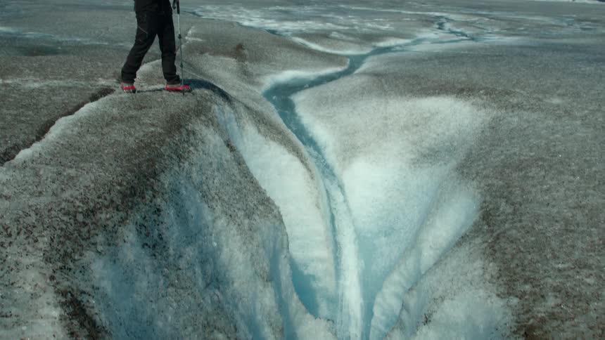A hiker stands at the edge of a deep crevasse on a glacier gazing down at the narrow stream of blue meltwater cutting through the ice