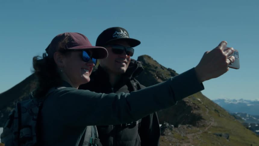 A couple takes a selfie together on a mountain trail under clear blue skies
