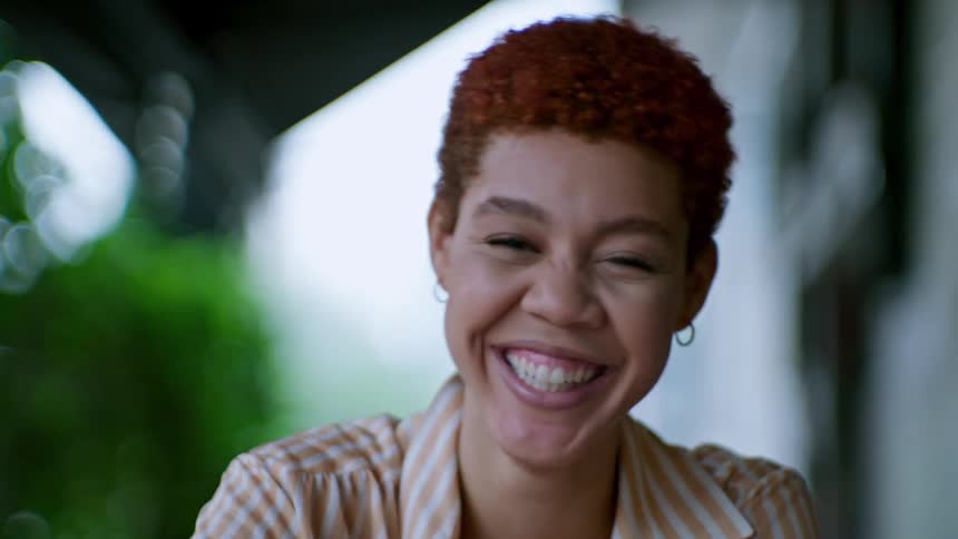 A person with short curly red hair and a striped shirt sits outdoors smiling and laughing