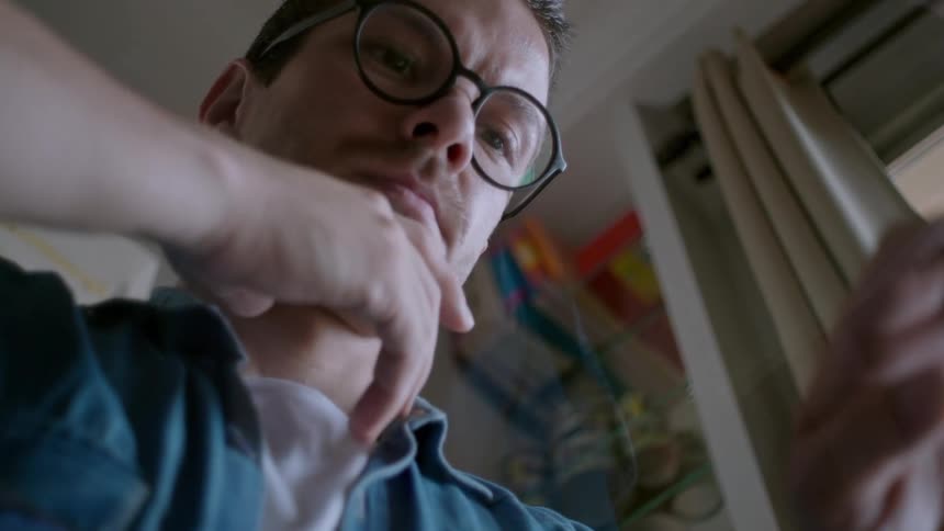 A man in round glasses and a denim shirt looks downward his hands raised midair as if miming a typing motion