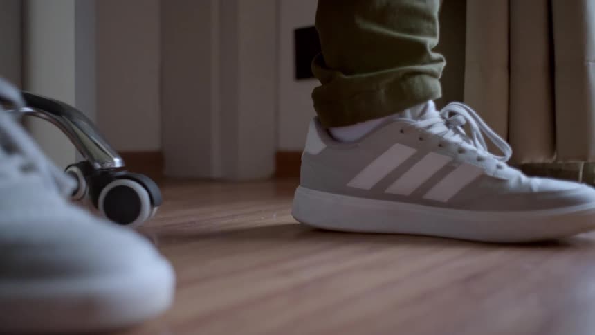 A person wearing light gray sneakers with white stripes and white socks steps on a wooden floor with one shoe prominently in the foreground and the other in midstep