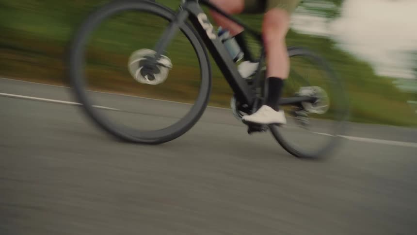 A cyclist wearing a green helmet rides along a paved road bordered by dense shrubs and tall leafy trees