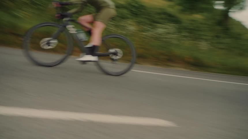 A female cyclist in dark athletic gear rides along a paved road a grassy hill rising on the right