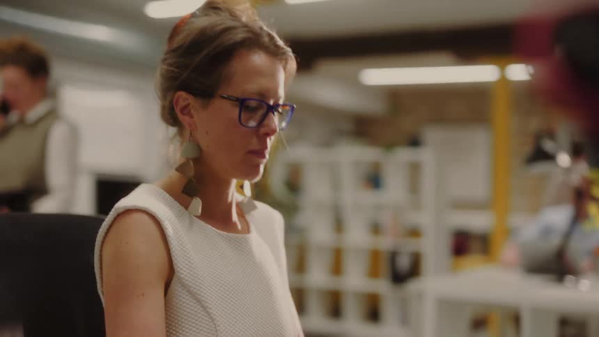 A woman with hair pulled back wearing a white sleeveless top and blue framed glasses works in a busy office space