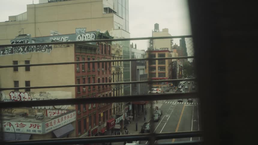 An urban street scene viewed through the bars of an elevated train or bridge features a mix of old brick buildings and modern glass structures with graffiti on walls