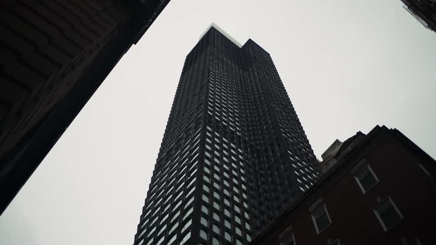 A towering dark skyscraper rises towards the sky flanked by other buildings that partially frame the view from below