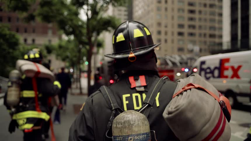 Firefighters in full gear with an fdny helmet walks away from the viewer carrying equipment on their back among a crowd of people and emergency vehicles on a city street