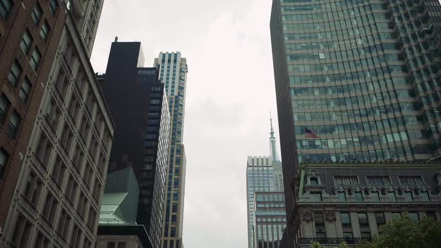 Tall buildings with varied architectural styles line a city street under an overcast sky with american flags visible on several structures