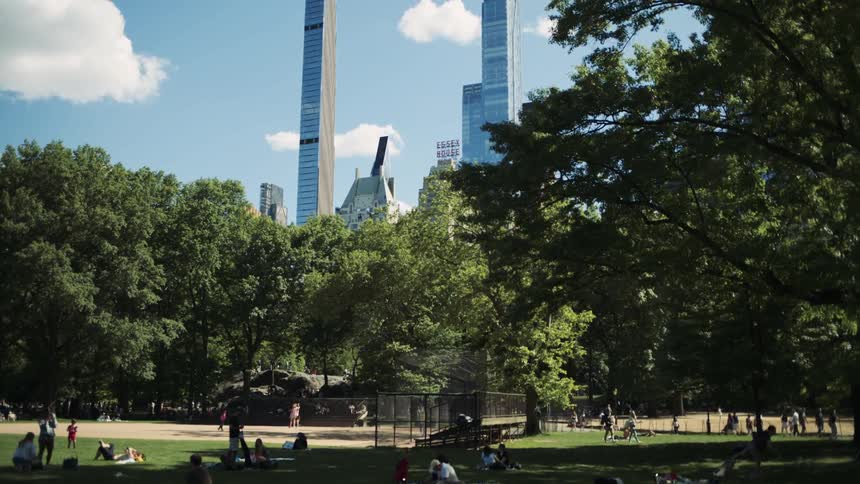 People relax and stroll and sit in a sunny park surrounded by skyscrapers