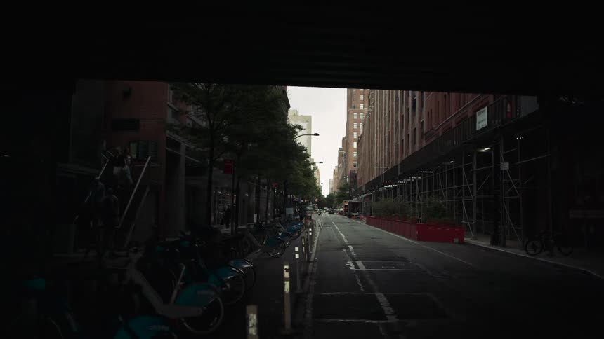 A street view from inside a moving vehicle revealing a dark tunnellike passage opening into a city street buildings line both sides of the road and parked bicycles are visible along the sidewalks