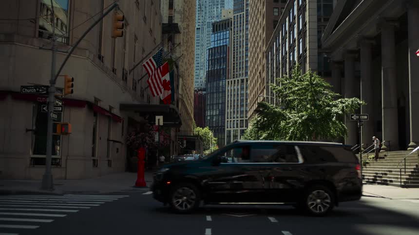 A busy city street lined with tall buildings and american flags shows cars a delivery truck and pedestrians under bright sunlight
