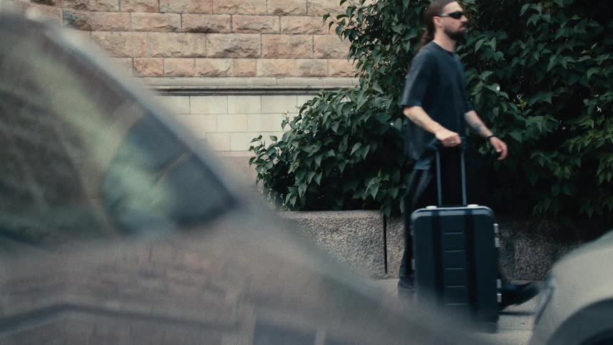 A man with long hair and sunglasses wearing a dark tshirt and pants walking on a sidewalk while pulling a dark suitcase he is walking past a building with stone walls and a green bush with parts of a car visible in the foreground