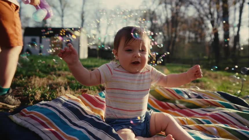 Baby having fun while sitting on a colorful rug on the grass and soap bubbles are flying around her