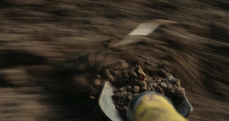 A closeup of a shovel coated in rich soil moving over garden earth with neatly planted green seedlings in the background