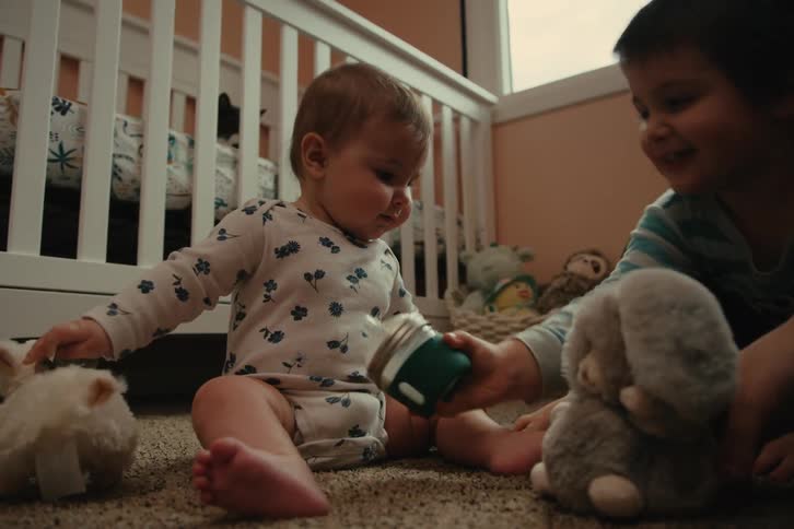 Young child helps baby drink from a bottle while sitting on the nursery floor