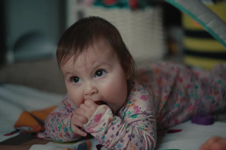 A baby in patterned pajamas chews on a toy while lying on the floor