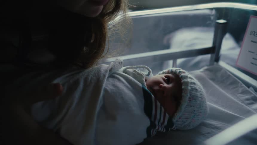 A newborn baby wrapped in a blanket lies in a hospital bassinet with a person leaning over the baby