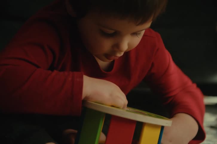 A toddler wearing a red shirt plays indoors with a toy drops a small red square into a hole in the toy and then tries to retrieve it