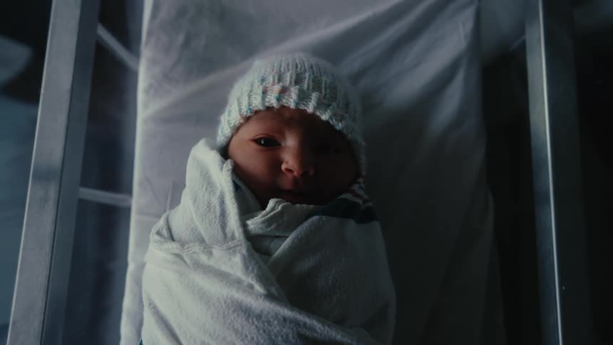 A newborn baby wrapped in a white blanket and wearing a light colored knit cap lies in a hospital bassinet