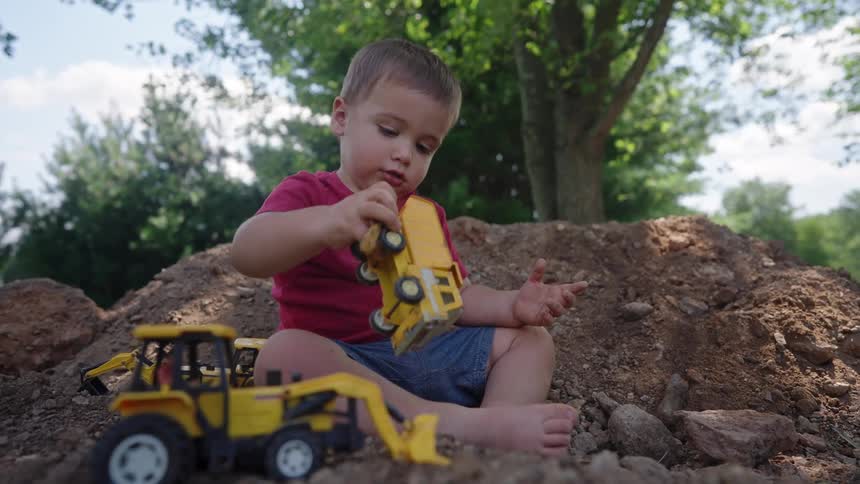 A young boy plays with toy construction trucks in a dirt pile outdoors
