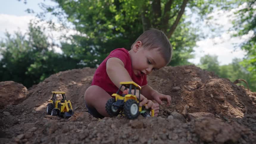Male toddler plays with toy vehicles on a soil heap outdoors during daytime