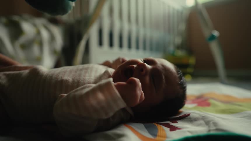 A baby lies on a colorful blanket near a crib looking up