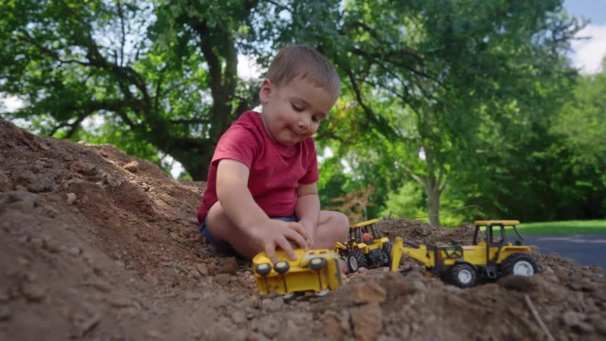 A toddler sits on the soil playing with a toy truck with two other bulldozer toys in front of him moving the toy around on the soil at midday