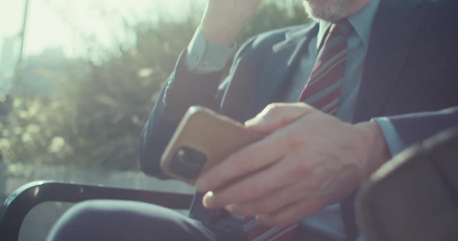 A man wearing a suit and red tie sits on a bench in the park at midday puts on his glasses and sets his phone aside