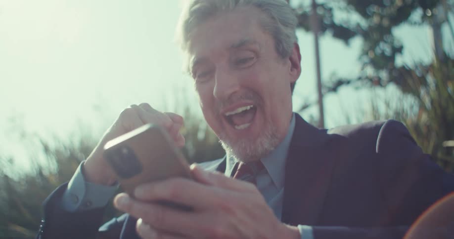 Man in suit and tie happily vibrating and interacting with smartphone while sitting in outdoor environment