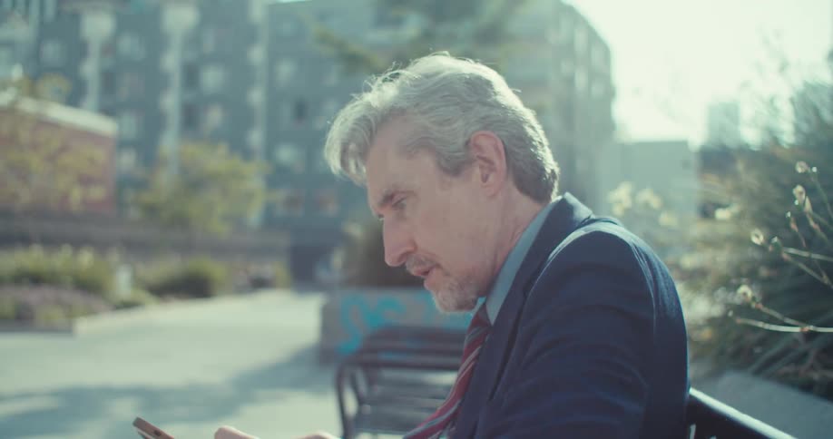 N older businessman sitting on a bench outdoors taking off his sunglasses and looking at his phone he appears to be focusing intensely on the screen reacting to something he sees