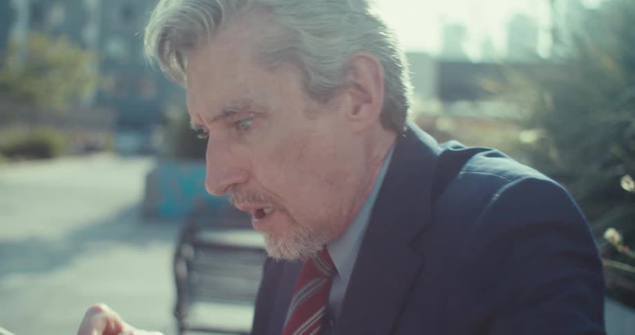 A businessman sitting on a city bench looks at his phone with growing shock and disbelief with the camera pushing in to capture his stunned reaction