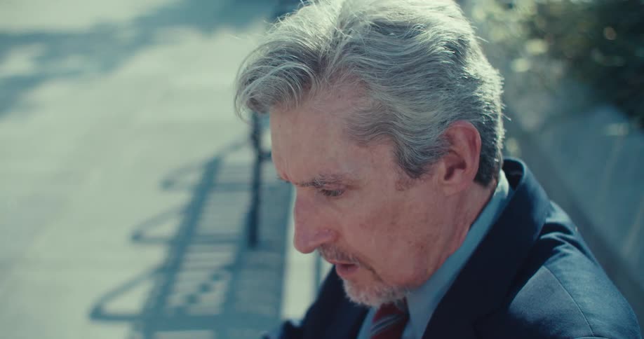 A man sitting on a park bench wearing sunglasses suddenly becomes shocked takes off his glasses and stares at a screen at midday