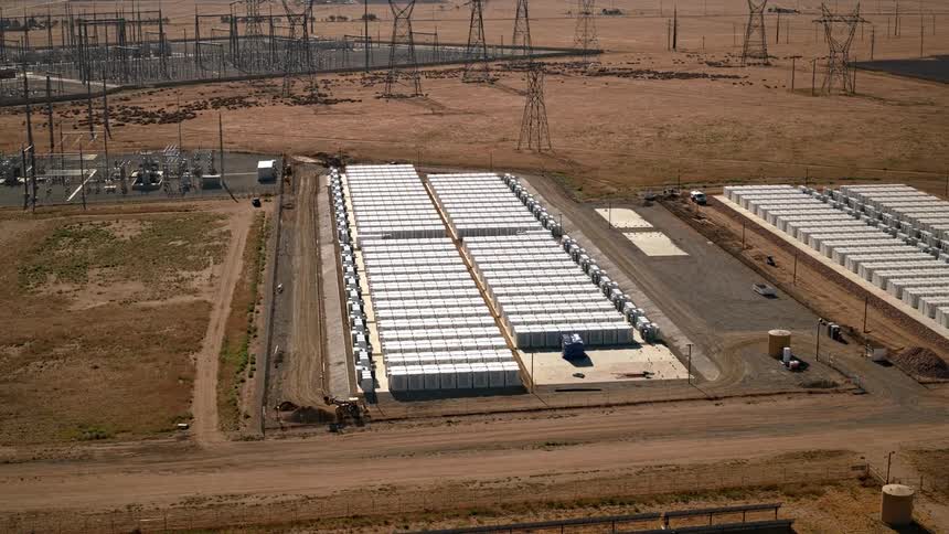 Rows of white battery units sit beside power lines in a dry open landscape with many towers in the background