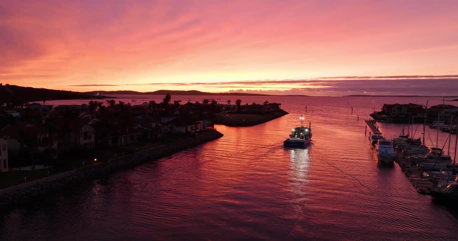 A boat sails through a narrow channel surrounded by docks and houses at sunset with the sky painted in vibrant shades of orange and pink