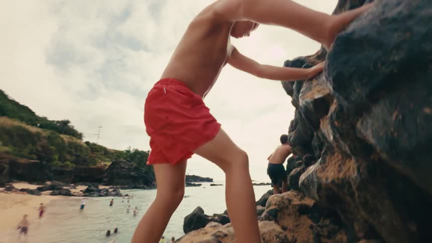 A boy in red shorts climbs a rocky formation near a beach