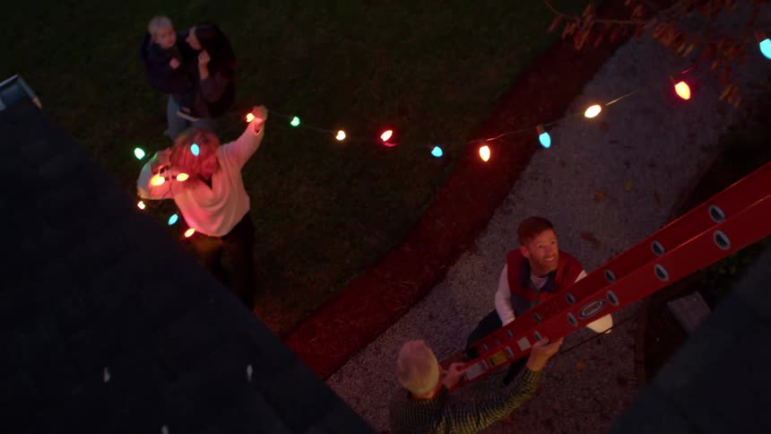 A family works together to hang colorful holiday lights at dusk